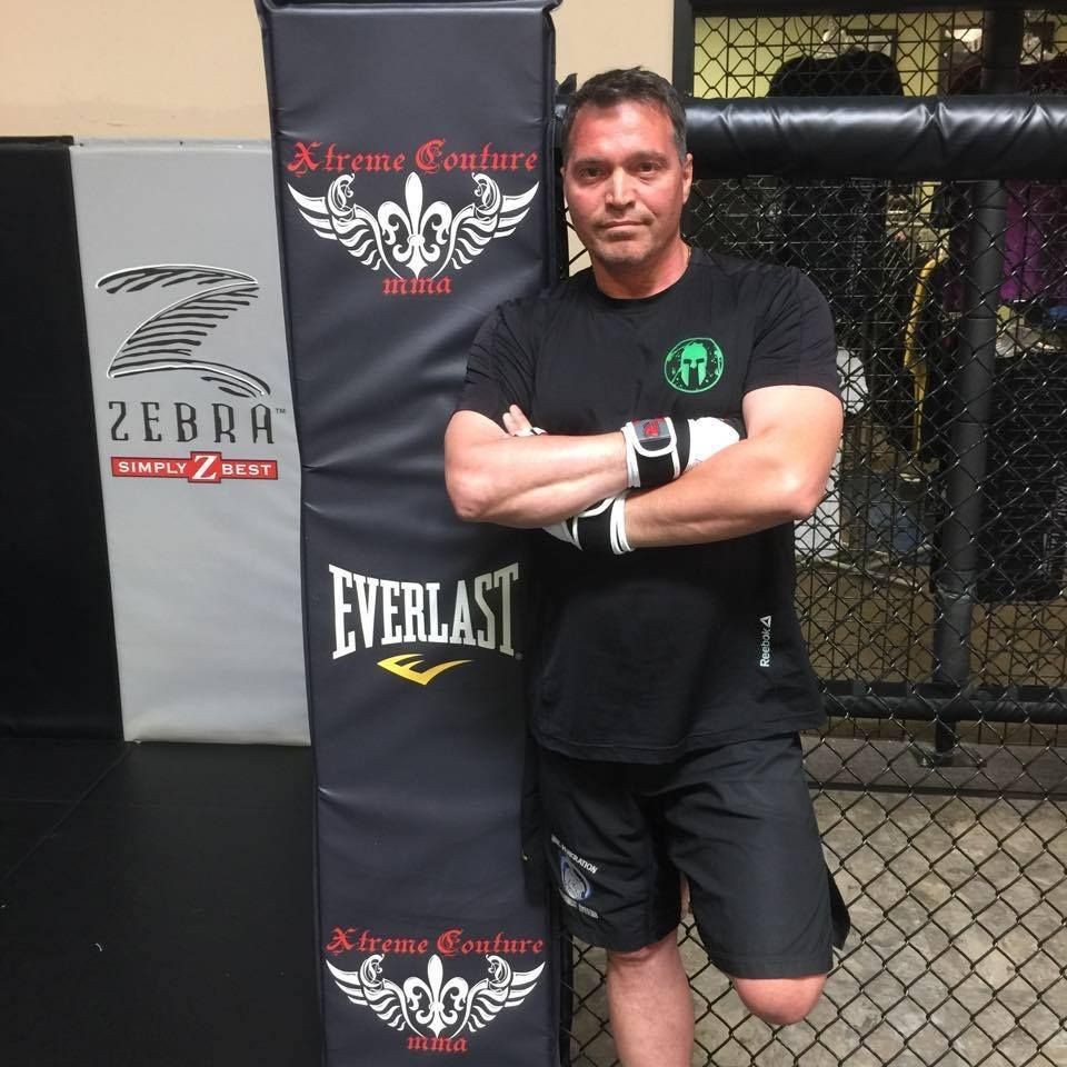 Man in a black workout outfit leaning against an Xtreme Couture MMA pad in a gym.